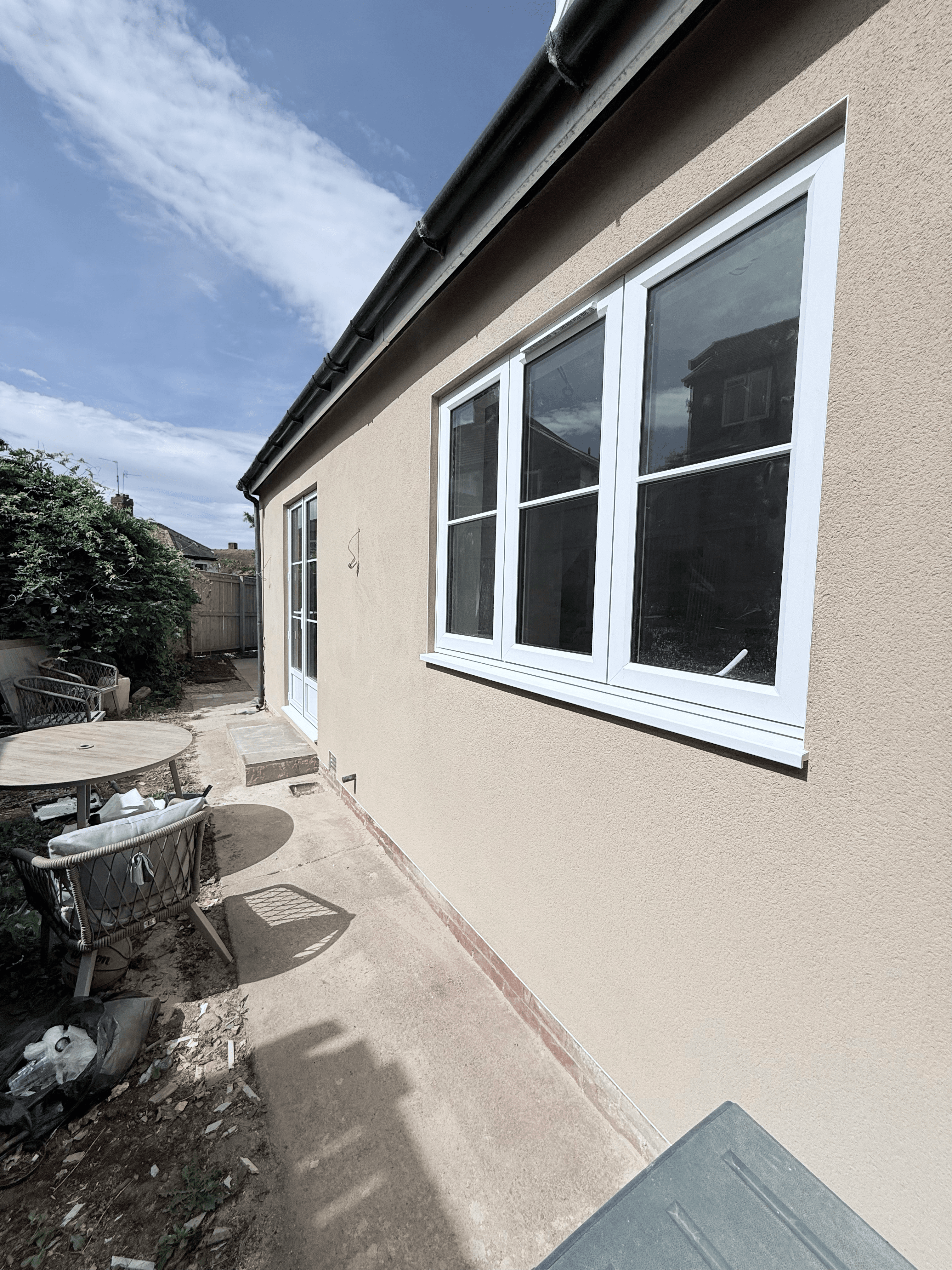 Tan exterior wall with a large white window and a patio area with outdoor furniture.