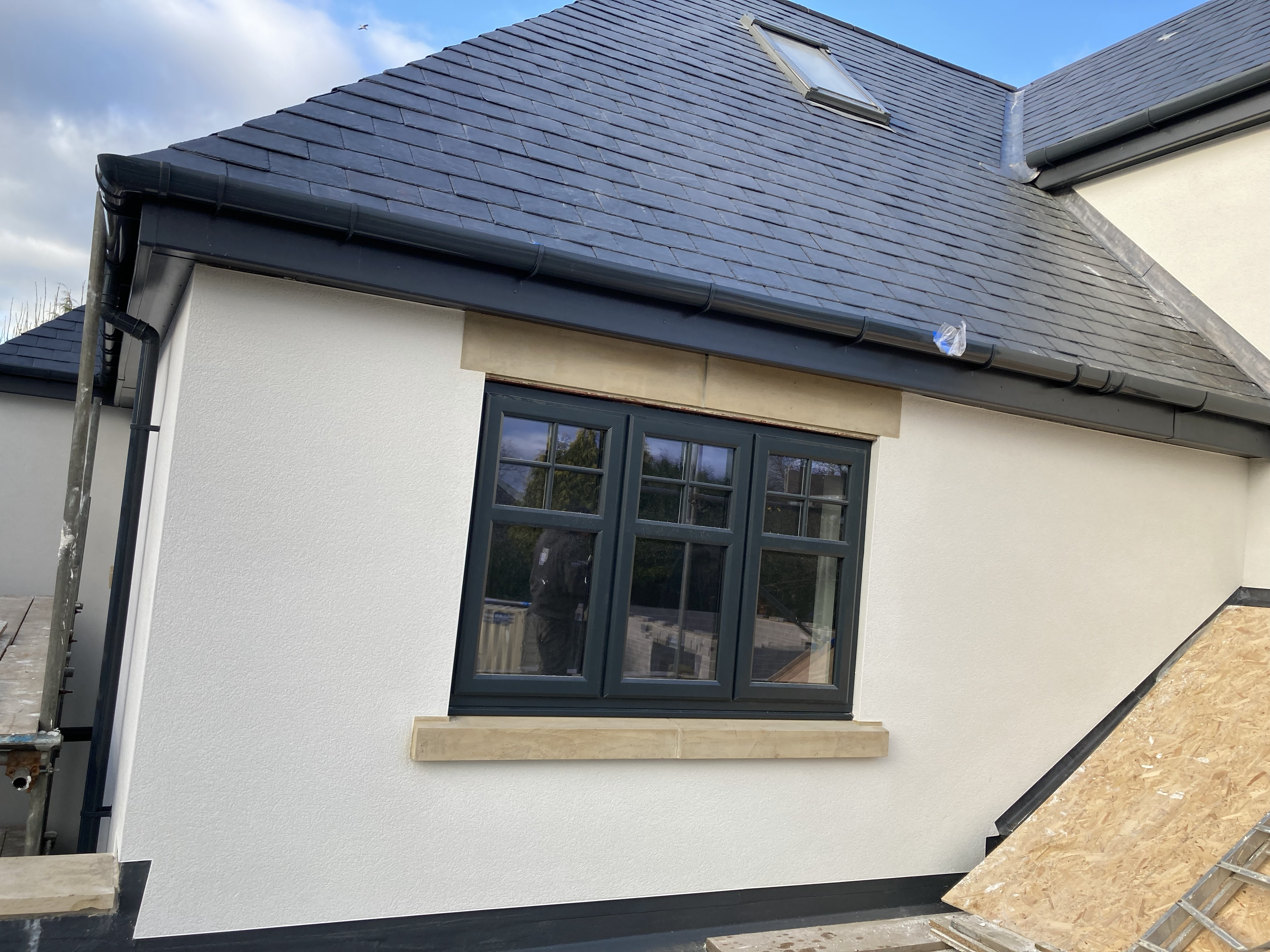 White rendered house wall with a dark grey window, stone sill, and slate tiled roof.