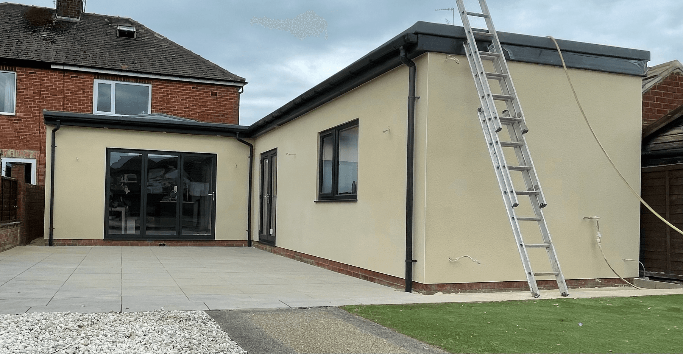Modern cream house extension with black window frames, stone patio, and a silver ladder.