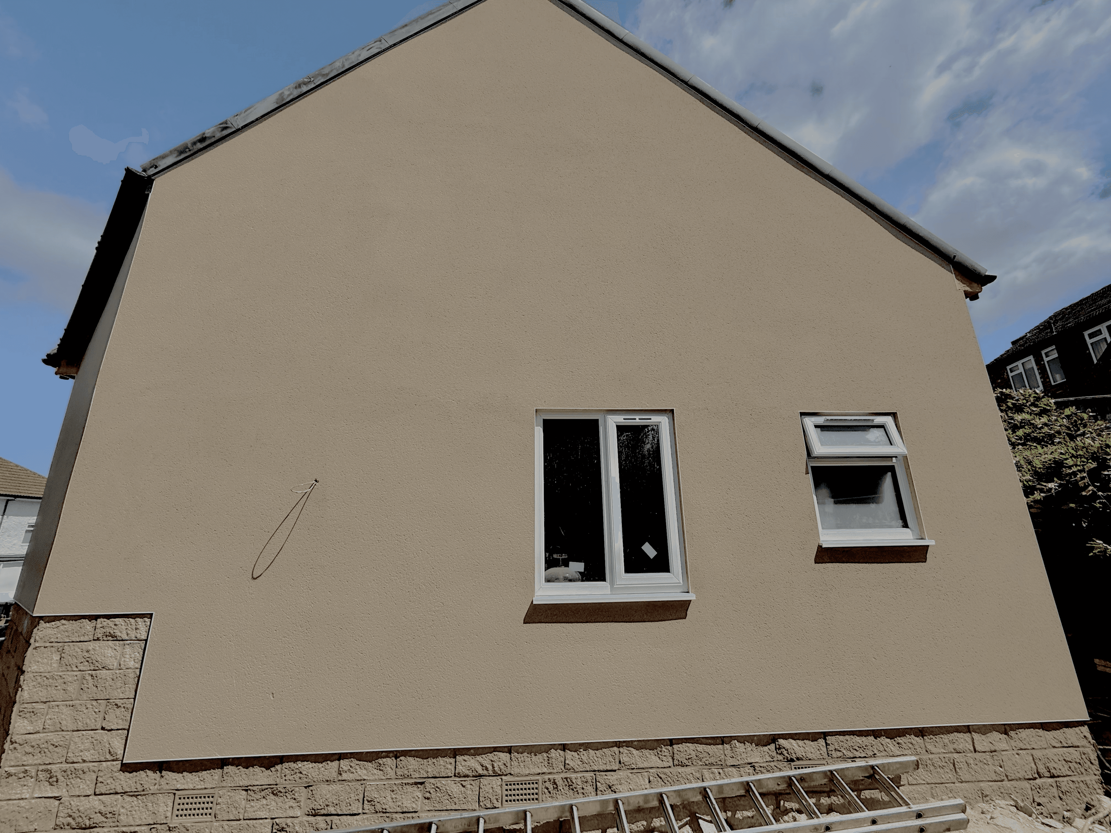 Beige rendered house wall with two white windows and a stone base against blue sky.