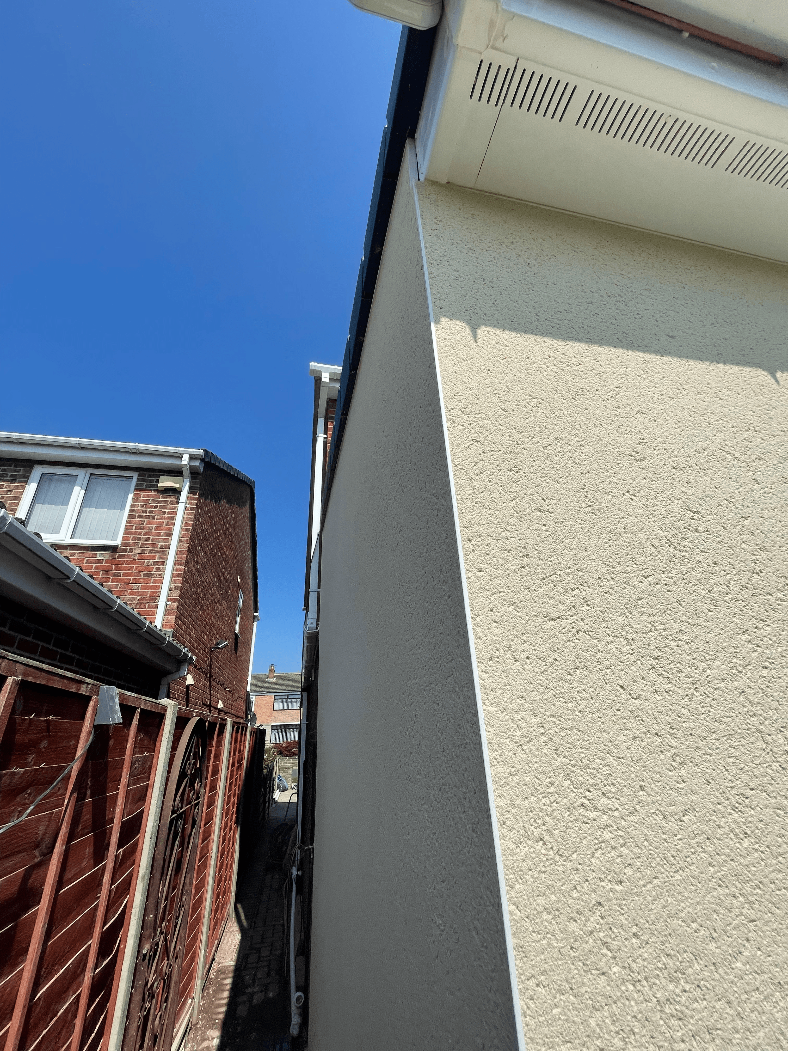 Narrow alleyway between a brick house and cream textured wall under a clear blue sky.